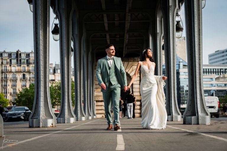 Couple de mariés marchant main dans la main sur le Pont Bir Hakeim à Paris, en plein soleil, lors d’un mariage marocain. La mariée sourit au soleil, la traîne sur le bras, pendant que le marié esquisse une grimace amusée.