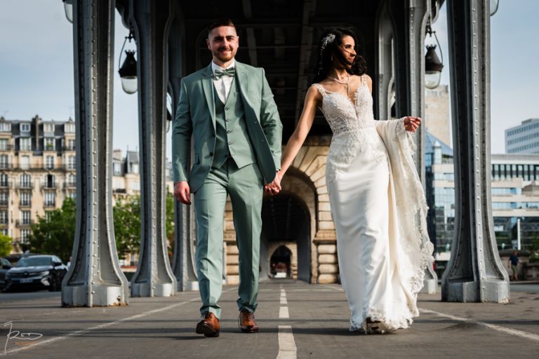 Marié regardant l’objectif en souriant tandis que la mariée offre toujours son visage au soleil, ses longs cheveux noirs légèrement emportés par le vent, sur le Pont Bir Hakeim à Paris, lors d’un mariage marocain.