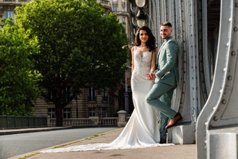 Couple de mariés marocains se tenant la main sur le Pont Bir Hakeim à Paris, regardant l'objectif, dans une scène épurée et lumineuse.