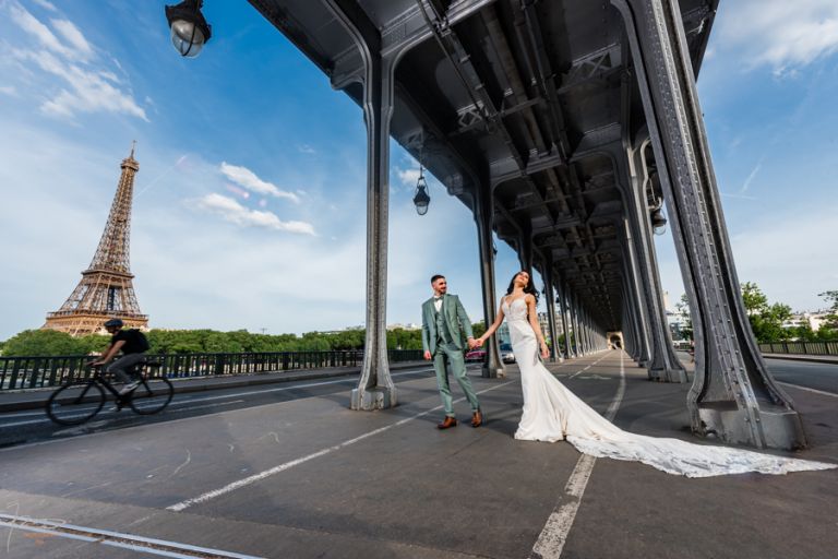Mariés marocains au centre du Pont Bir Hakeim à Paris, avec la Tour Eiffel à gauche et le ciel bleu à droite, photographiés en grand angle à 12mm. Le marié tire doucement la main de la mariée qui se laisse emmener.