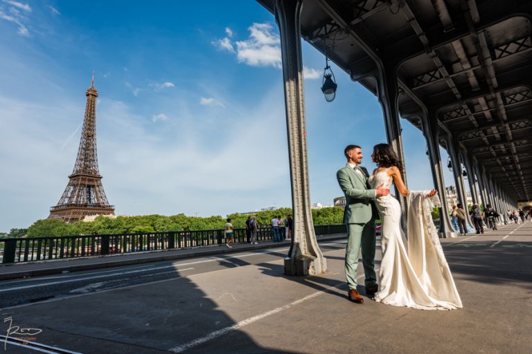 Mariés marocains enlacés sous le Pont Bir Hakeim à Paris avec la Tour Eiffel en arrière-plan, la mariée tend sa traîne dans une pose élégante.