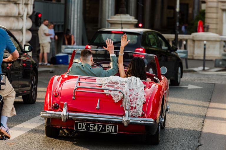 Mariés marocains quittant la scène en voiture de collection, montrant leurs alliances, voile de la mariée posé sur le coffre, à Paris.