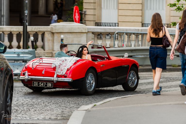 Voiture de mariage tournant à droite dans les rues de Paris, la mariée regarde le photographe en souriant, effet de flou artistique sur les roues en mouvement.